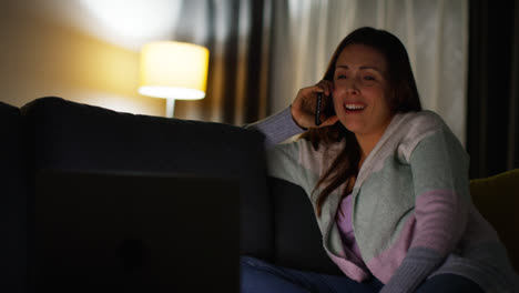 Smiling-Woman-Sitting-On-Sofa-At-Home-At-Night-Talking-On-Mobile-Phone-And-Watching-Movie-Or-Show-On-Laptop-1